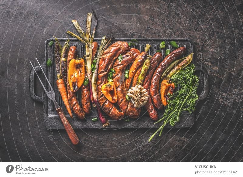 Various grilled meat sausages with roasted vegetables on cast iron grill pan with meat fork on dark rustic background. Grill party Oktoberfest German food. Top view. Traditional cuisine