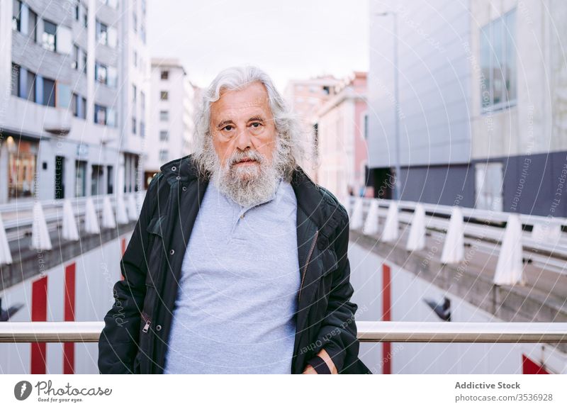 Senior male traveler standing on bridge with urban street buildings behind senior man explore serious tourism beard gray hair lean metal railing modern downtown