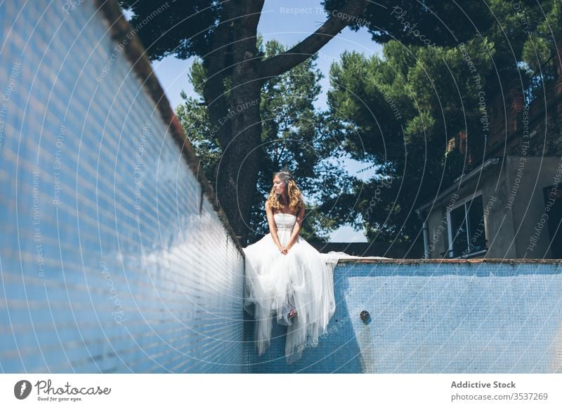 Pensive young bride in white wedding dress resting on edge of empty swimming pool woman elegant style garden party tree sensual dream summer romantic green