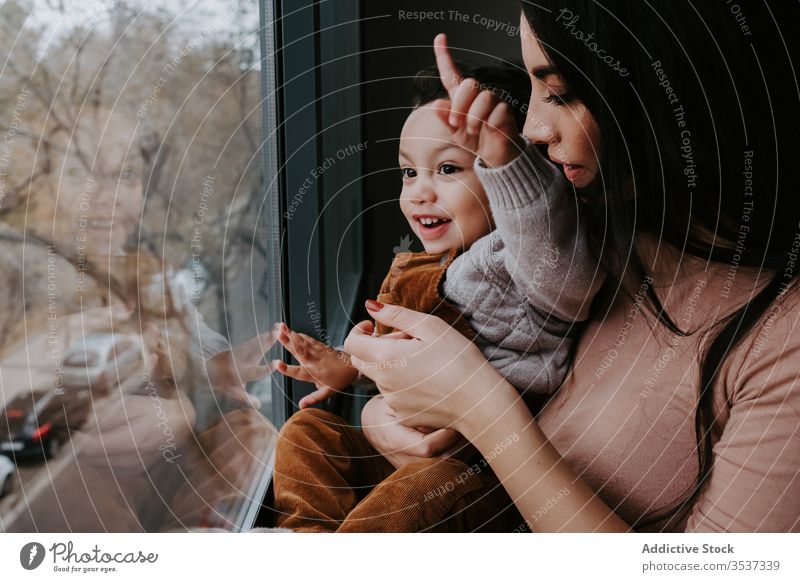 Positive young mother and little son standing near window and admiring view home curious explore point content excited cute child smile embrace kid parent