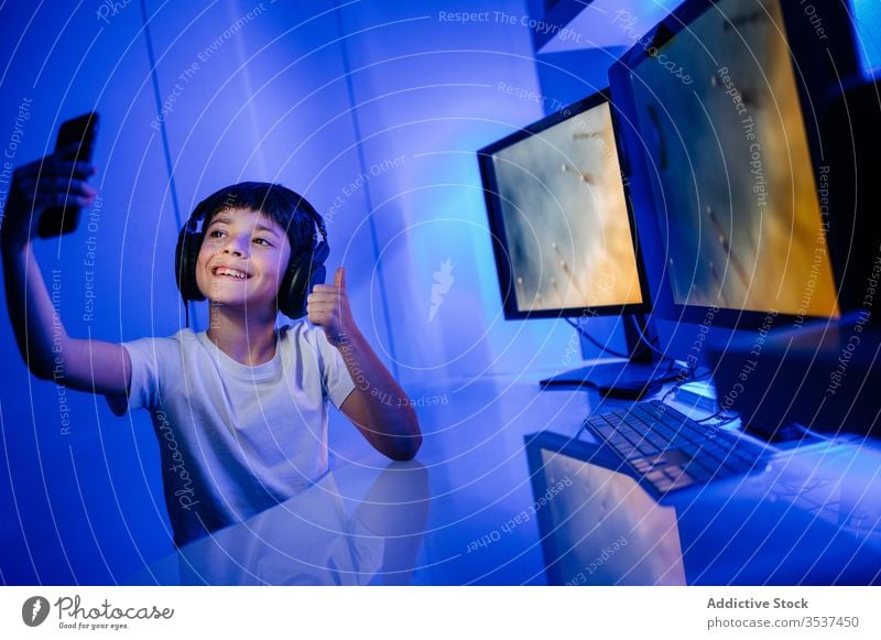 Cheerful boy in headset taking selfie on smartphone at home headphones thumb up toothy smile desktop computer using gadget device mobile phone cellphone monitor