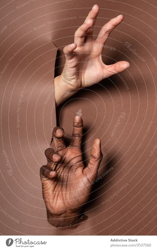 Anonymous crop multiracial couple bursting through paper curtain hand hole show together finger joy leisure fun arm palm relax positive people rest torn detail