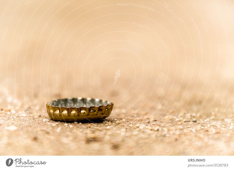 A crown cork on sandy soil rarely remains alone Crown cork beer closure Ground jettisoned Lie Gold Beige Bottle lid close up Close-up Deserted Sheet metal piece