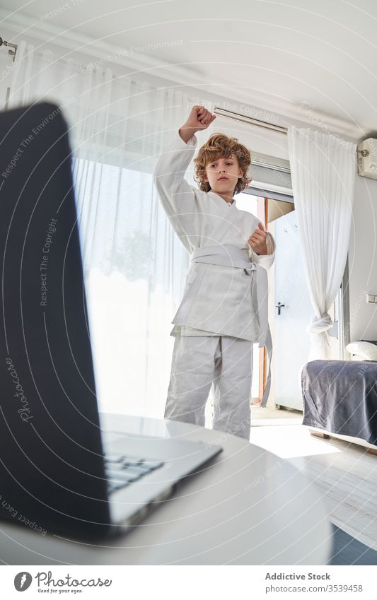 Young boy in white kimono learning judo on laptop at home young online tutorial position practice training childhood activity curly hair computer netbook