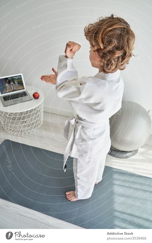 Young boy in white kimono learning judo on laptop at home young online tutorial position practice training childhood activity curly hair computer netbook