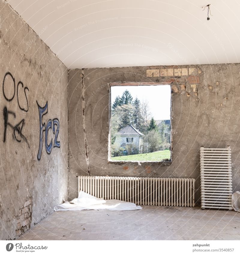 Room without furniture and without wall cladding, brickwork peeps out through plaster, two radiators leaning against the wall, graffiti on the walls, through frameless window opening views of meadow and next villa-like house