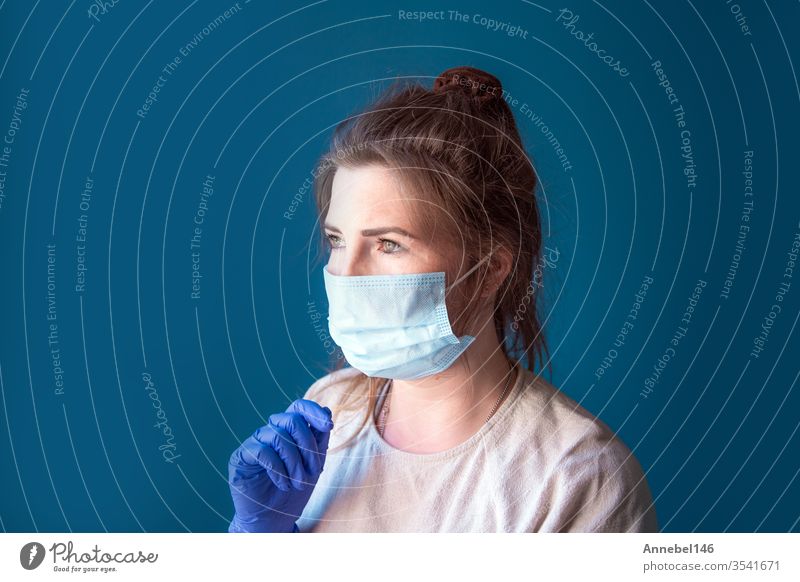 Young woman wearing protective gloves and face mask inside a home in quarantine looking bored and sad, for Covid-19 Coronavirus, with blue background medical