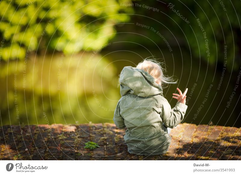 Rear view of a girl - out in nature sits Blonde Child Toddler sedentary Water green huts Anonymous hand movement Playing self-employment on one's own by oneself