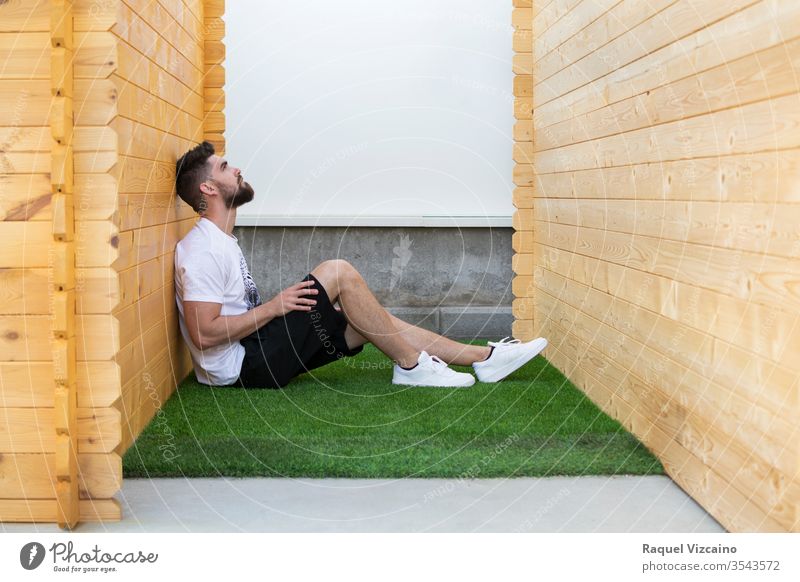 A handsome man sitting on the artificial grass between two wooden cabins. seated white health relaxation wellness house home zen outdoors relaxing loneliness