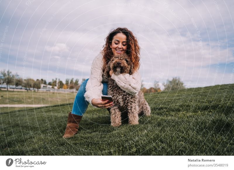 A beautiful woman is in the meadow with her dog. The owner is hugging her pet while trying to take a photo with her phone. The pet is a Spanish water dog with brown fur.