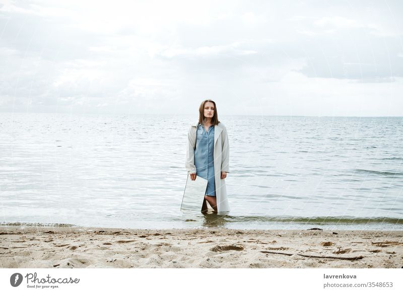 Closeup of woman standind on a shore in blue dress holding a mirror with water reflecting in it, selective adult beach caucasian clean coast concept environment