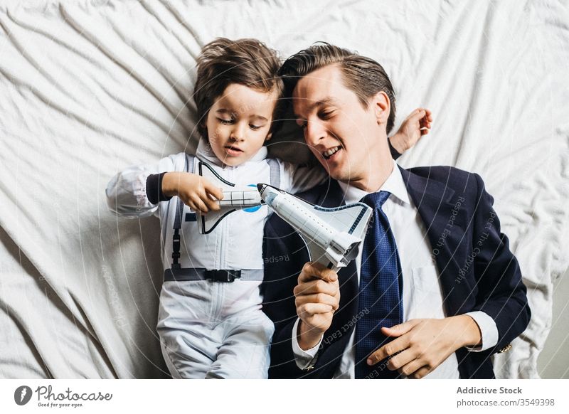 Content ethnic son and father lying on bed together astronaut costume boy dad play toy having fun diverse multiracial spacesuit parent spaceship smile kid