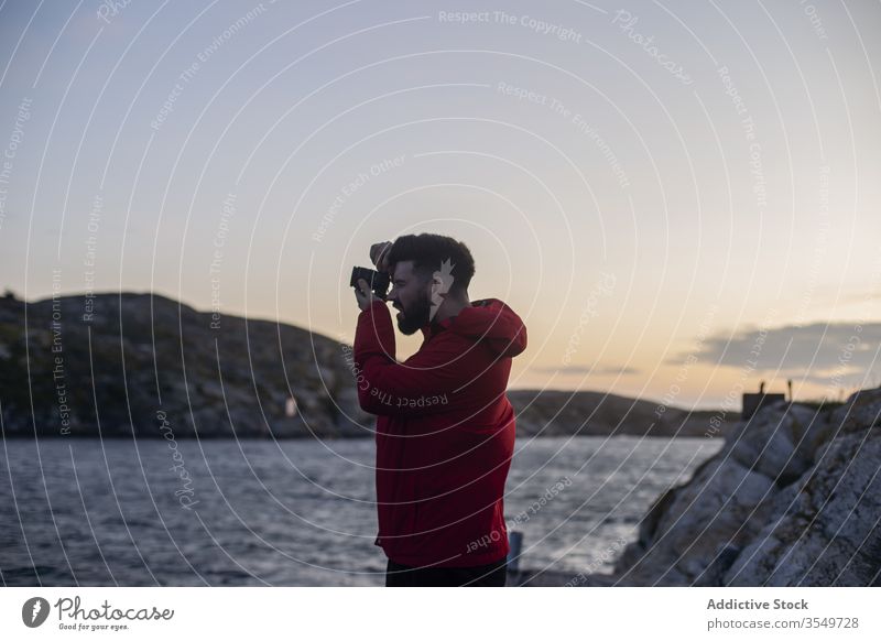 Man taking picture on photo camera on sea shore man take photo rocky photographer coast cliff sundown evening nature trip travel hoodie wanderlust adventure