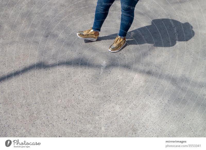 Chubby female wearing  tight blue jeans and golden white fashion sneakers waiting on a sidewalk with shadow and tree branch shadow slightly overweight woman