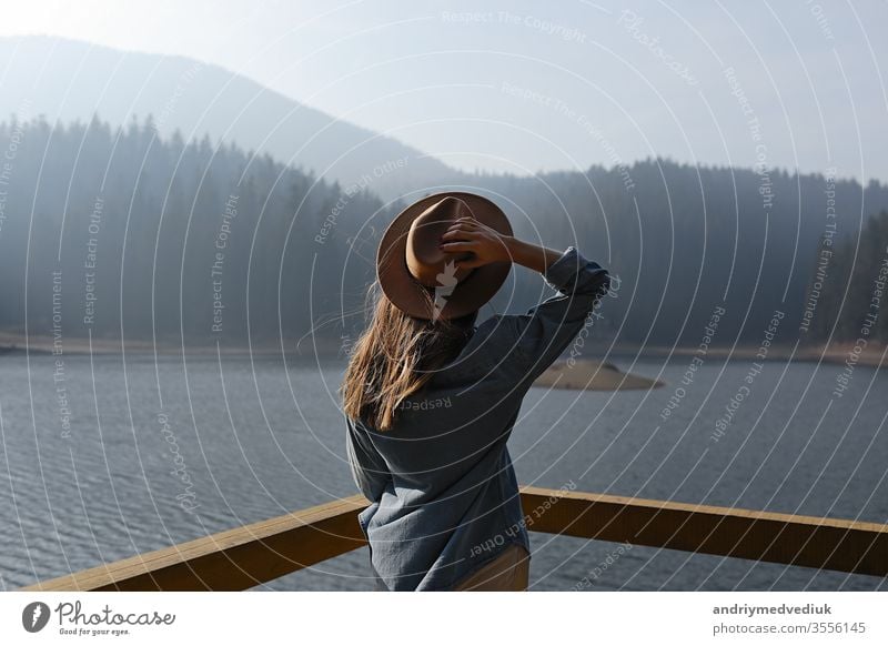 happy young woman in hat enjoys lake view in mountains . Relaxing moments in forest. Back view of stylish girl enjoys the freshness outdoor. Freedom, people, lifestyle, travel and vacation