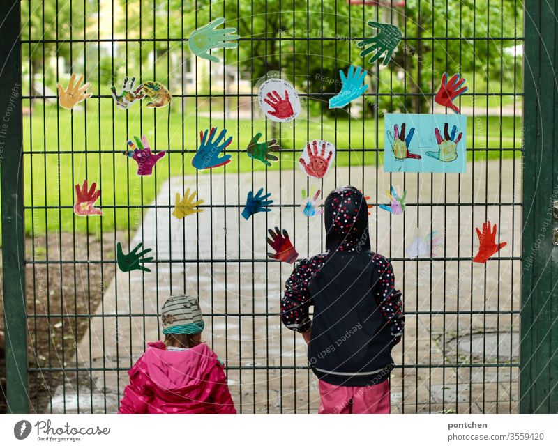 Two small female children in rainwear stand in front of the garden gate of a kindergarten and look inside. Colourful handicrafts hang from the gate. Longing, missing. Closure