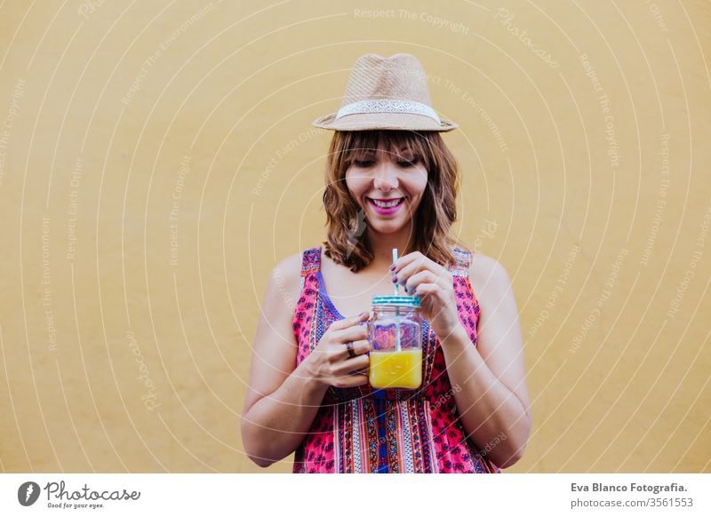 portrait of young caucasian woman outdoors over yellow background enjoying a cup of healthy orange juice. summer time city lifestyle curvy real people real life