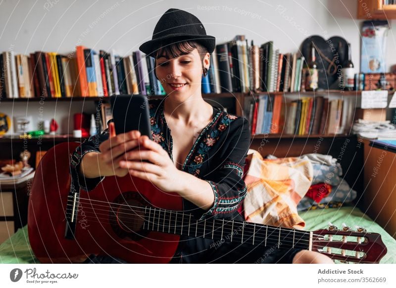 Cheerful woman with guitar browsing smartphone at home acoustic trendy message using cellphone music female weekend style wear hat bed sit relax gadget device