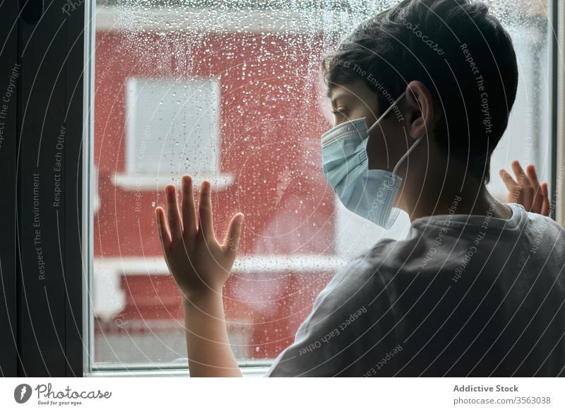 Sad boy in medical mask near window coronavirus epidemic home sad unhappy self isolation social distancing child kid face mask stand lean observe rain street