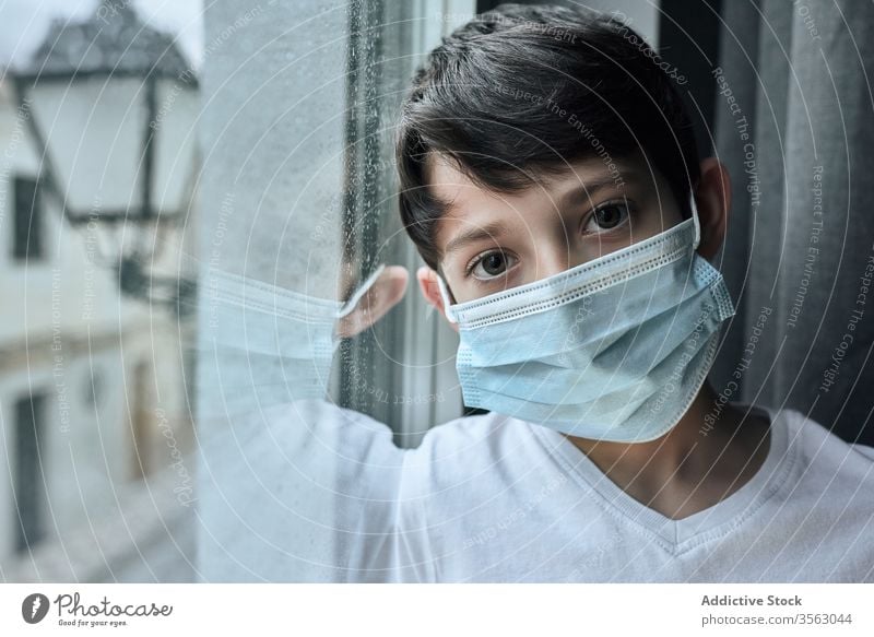 Sad boy in medical mask near window coronavirus epidemic home sad unhappy self isolation social distancing child kid face mask stand lean observe rain street