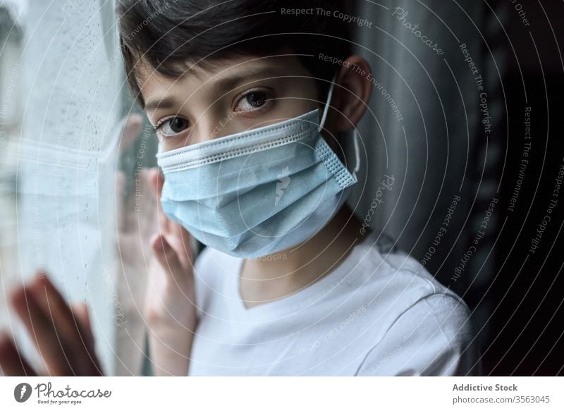 Sad boy in medical mask near window coronavirus epidemic home sad unhappy self isolation social distancing child kid face mask stand lean observe rain street