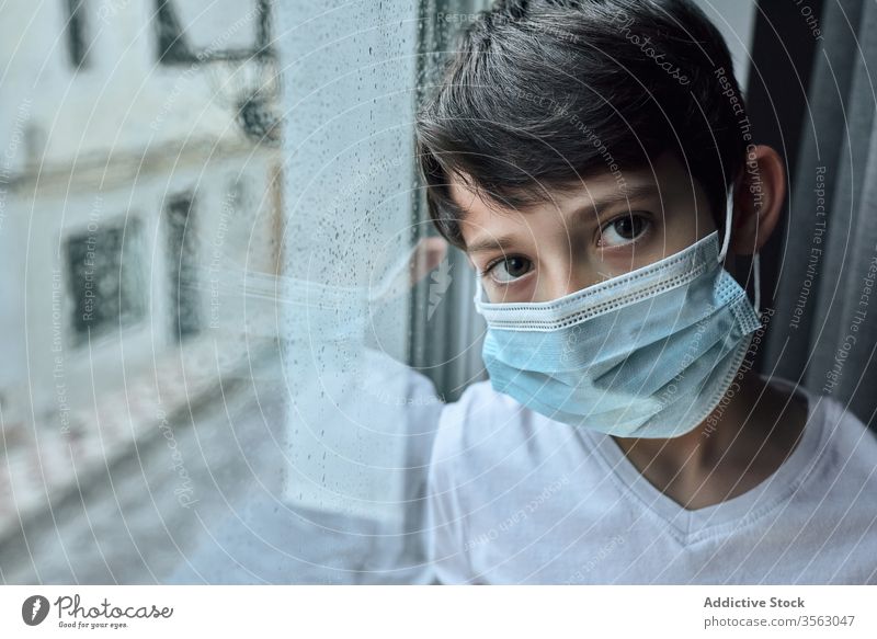 Sad boy in medical mask near window coronavirus epidemic home sad unhappy self isolation social distancing child kid face mask stand lean observe rain street