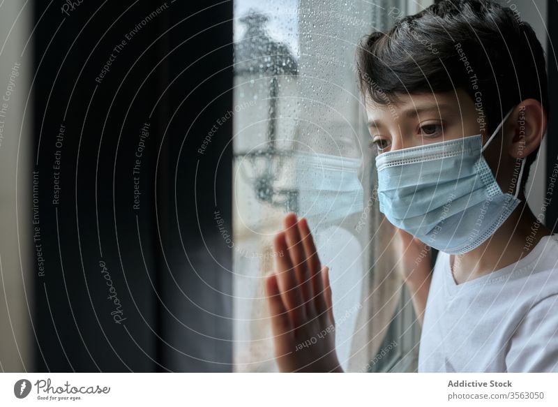 Sad boy in medical mask near window coronavirus epidemic home sad unhappy self isolation social distancing child kid face mask stand lean observe rain street