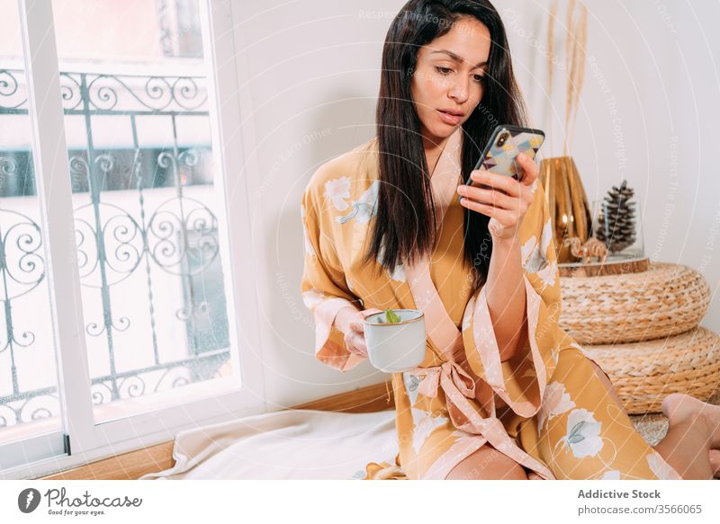 Attractive young woman browsing smartphone while having a tea mat chill home robe zen sensual social media texting weekend floor sleepwear communicate internet