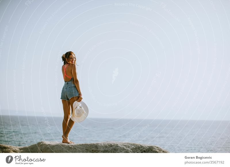 Young woman in bikini standing on rocks by the sea adult attractive beach beautiful blue brunette caucasian coast day female greece hair holiday leisure