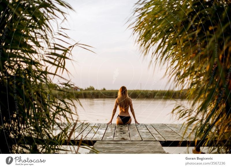 Young woman in bikini sitting on a pier at the lake alone attractive beach beautiful beauty blonde carefree caucasian female model nature outdoor person posing