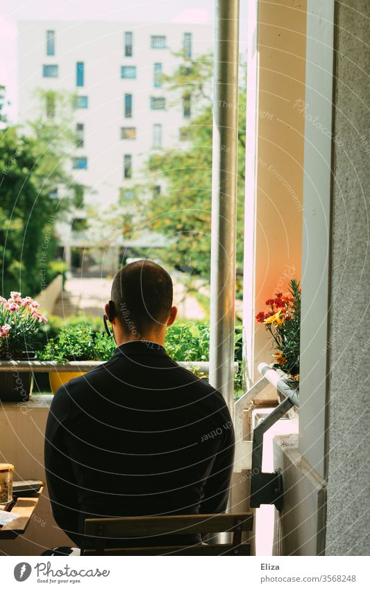 Man in black clothes with headset in his ear sits on the balcony in the sun and looks outside Balcony Sun good weather sunshine Rear view planted flowers Sit