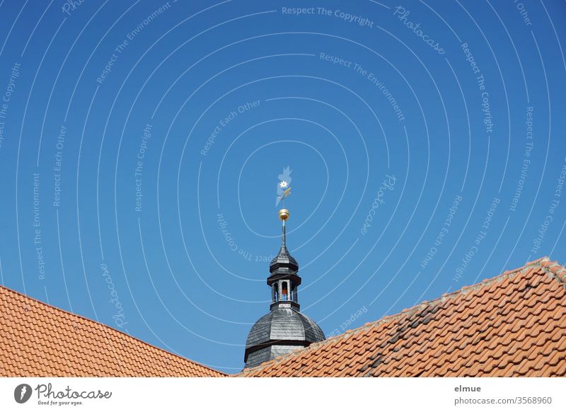 The top of a church tower looks out from behind red tiled roofs Church spire Tiled roof Roof Beautiful weather Weathercock Roofing tile Blue sky Red