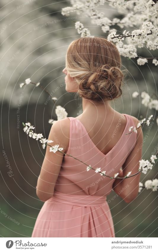 young stylish woman near flowering tree in spring park. blonde girl with hairstyle in pink dress backdrop. selective focus people blossom fashion luxury cherry