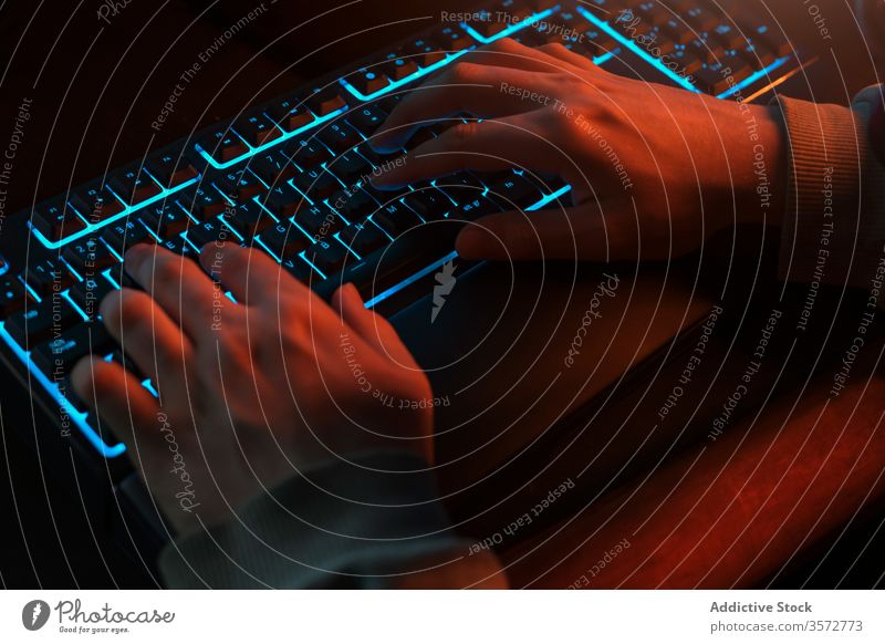 Crop programmer typing on keyboard with blue backlight in dark room at home night hand man comfort ray chat hacker freelance search browsing surfing gamer job