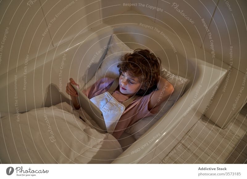 Cheerful boy using tablet in bathroom cartoon bathtub child cozy pillow fun lying watch weekend cheerful rest kid relax gadget device browsing curly hair