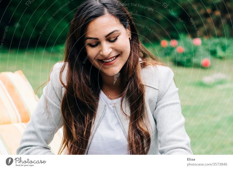 Brown-haired young pretty woman sitting on a deck chair in the garden and looking down with a shy smile Woman Garden out Sit Long-haired brown-haired brunette