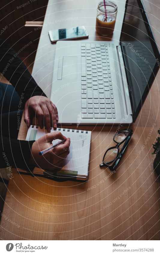 Female hands taking notes on her pad. Room with laptop, mobile and glasses. Indoor plant on a light wooden table with wooden wall. Working from home for quarantine.