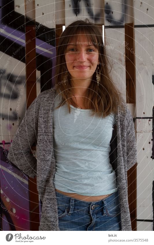 Pretty, cheerful, grinning young people in front view, with long, brown hair and earrings, standing leaning, belly-free, grinning, in front of an old, painted wall with graffiti and looking smiling, smiling and carefree into the camera.