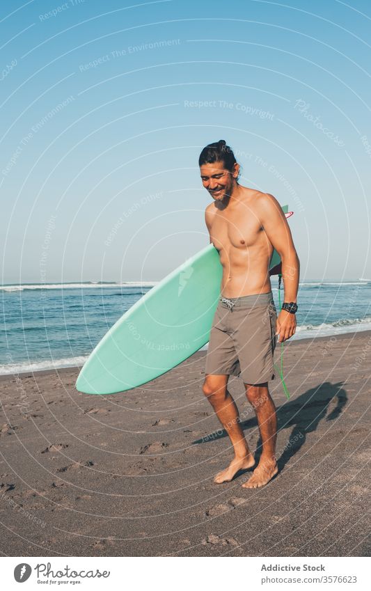 Cheerful surfer with surfboard standing on beach man sea happy active ocean coast smile shirtless ethnic male lifestyle sand summer shore vacation water