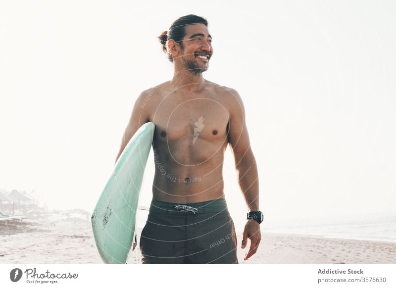 Cheerful surfer with surfboard standing on beach man sea happy active ocean coast smile shirtless ethnic male lifestyle sand summer shore vacation water