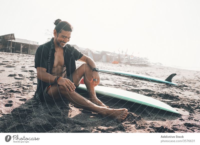 Happy ethnic man with surfboard sitting on beach happy positive rest sea enjoy sand summer young hispanic male lifestyle vacation coast ocean shore journey glad