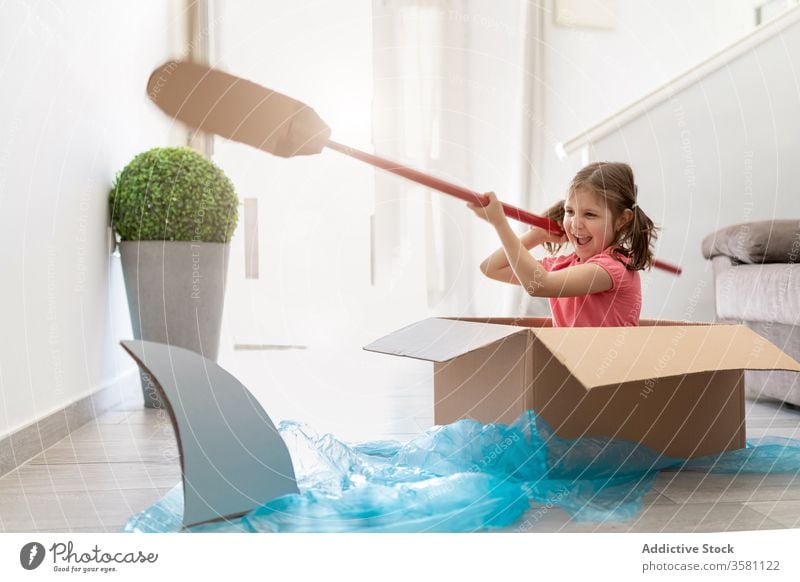 Joyful small girl in carton box sailing away from shark boat paddle game imagination fun playful kid sailor home child having fun joy pretend float childhood