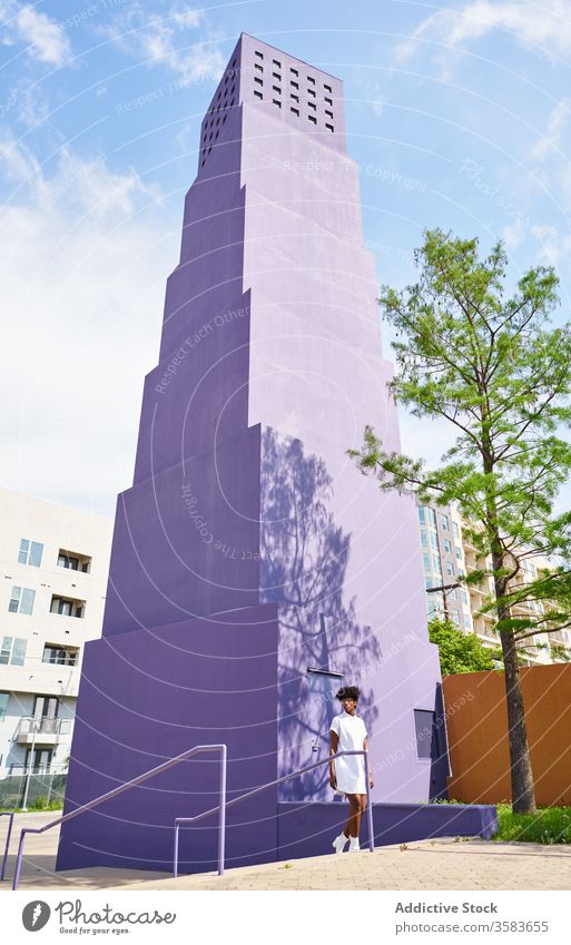 Stylish black woman walking on street against purple high rise building in downtown dress fashion trendy outfit female architecture style streetlamp urban