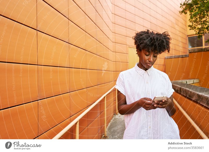 Focused black woman using smartphone on street against vivid exterior of modern building messaging pathway chill browsing smile social media facade surfing read