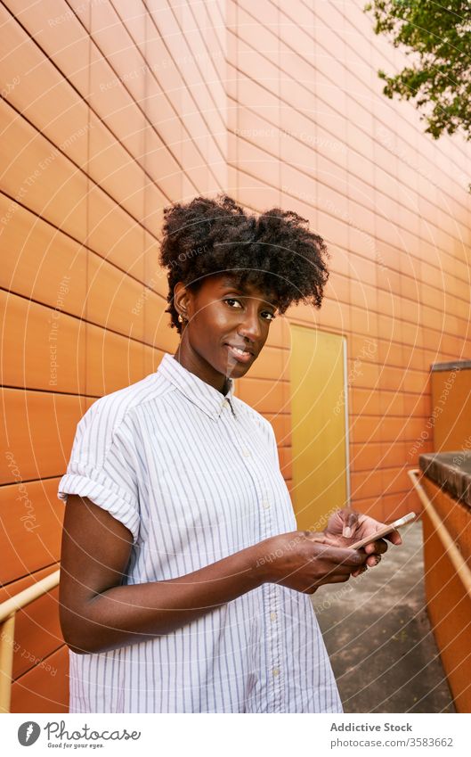 Happy black woman using smartphone on street against vivid exterior of modern building messaging pathway chill browsing use smile social media facade surfing