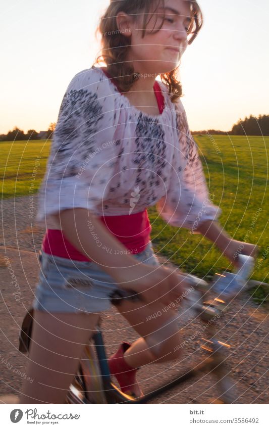 Smiling, the brunette teenager, in a fashionable blouse and shorts, pedals on the pedals, grinning cheekily. Summery, fast, dynamic bike ride on the road, past green grass of a meadow and trees, with blurred by the movement.