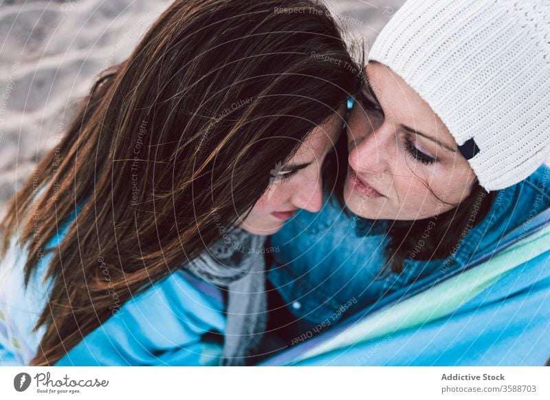 Positive lesbian couple resting on beach content sea cold cuddle season warm clothes sand lgbt spend time women gay young casual outfit hat jeans jacket shore