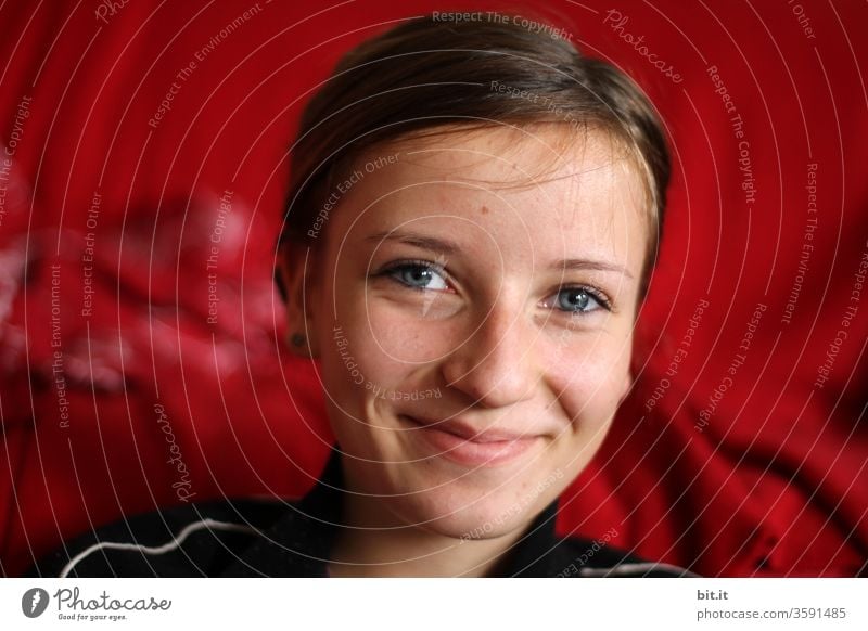 Happy, optimistic, handsome teenager smiles friendly, mischievously, in front of a red background into the camera. Fun-loving, vivacious, happy teenager with blue eyes, beaming eyes, post in front of red, glowing curtain, in the theatre.