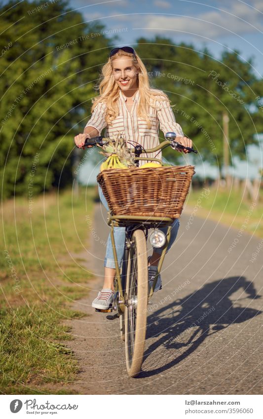 Happy woman smiling while riding a bicycle with a basket full of fresh and healthy fruits in a sunny day of summer in the countryside happy rural track meadows