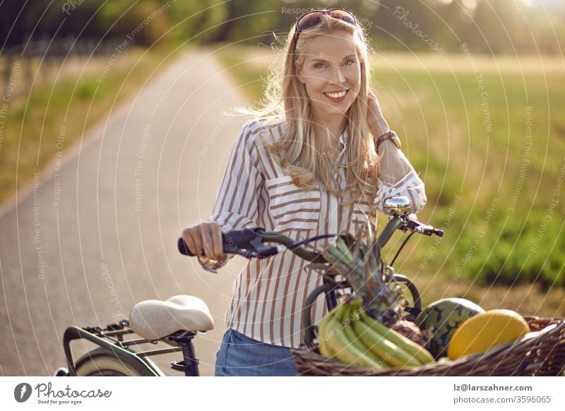 Woman using her bicycle to buy fresh produce standing on a rural road backlit by a warm glow of the sun smiling happily at the camera as she holds her bike with basket full of fruit and vegetables